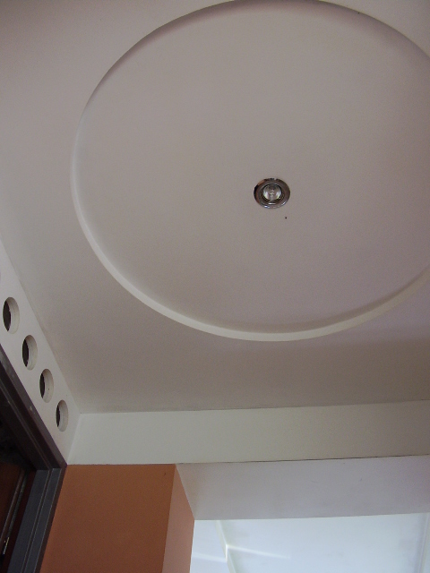 Biały sufit z okrągłym, podwieszanym elementem i wbudowanym oświetleniem punktowym, widok z dołu, fragment ściany z otworami wentylacyjnymi.